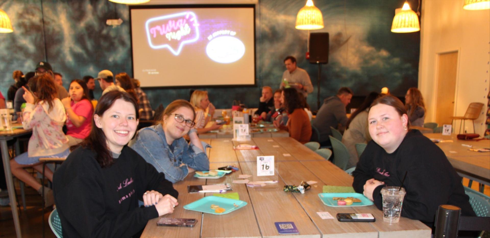 Three women sitting at a table at the GOTR NOVA Trivia Night event with the Trivia Night logo on a screen in the background.