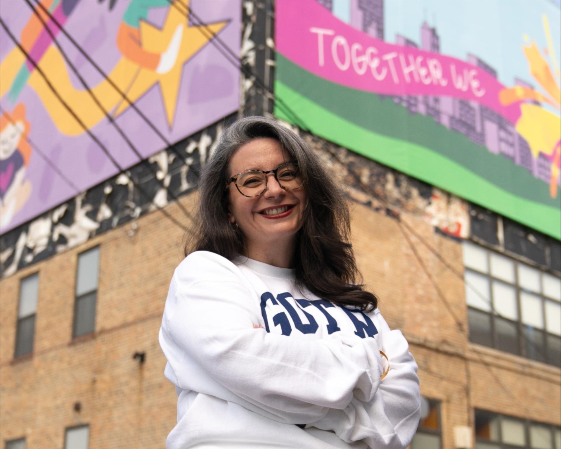 GOTRC Executive Director Jill Eid, wearing a white GOTR sweater, smiling standing in front of the GOTRC mural